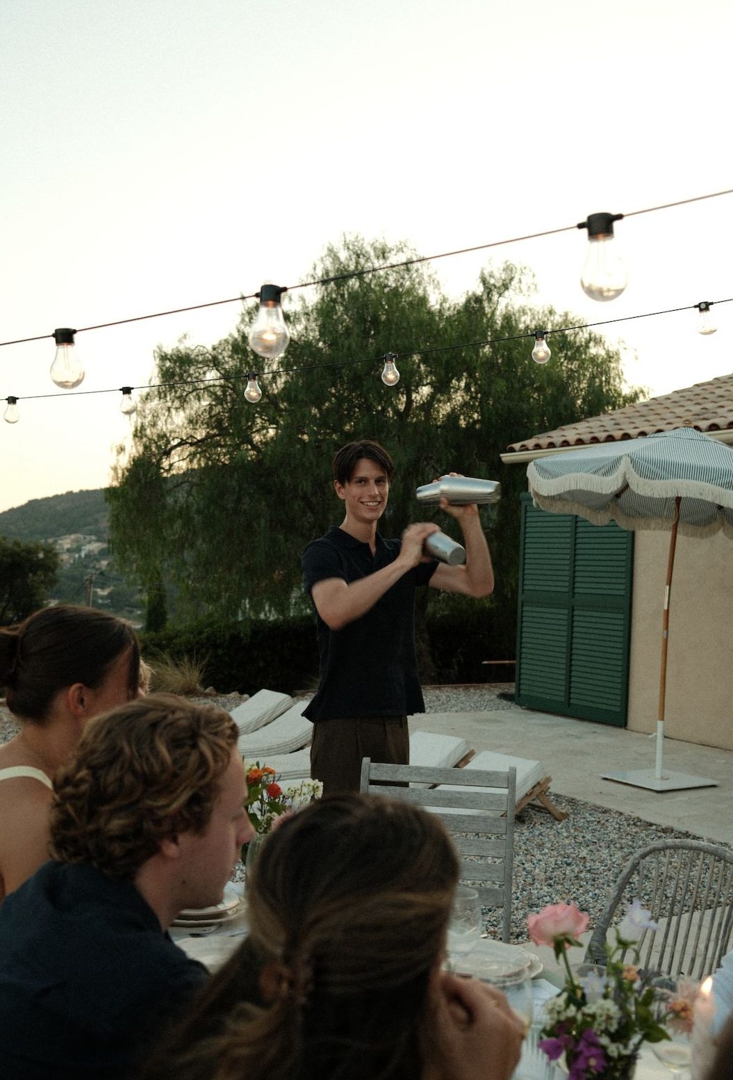 A host shaking cocktails at dusk on a Provence terrace, string lights overhead, with guests seated at the table in the foreground