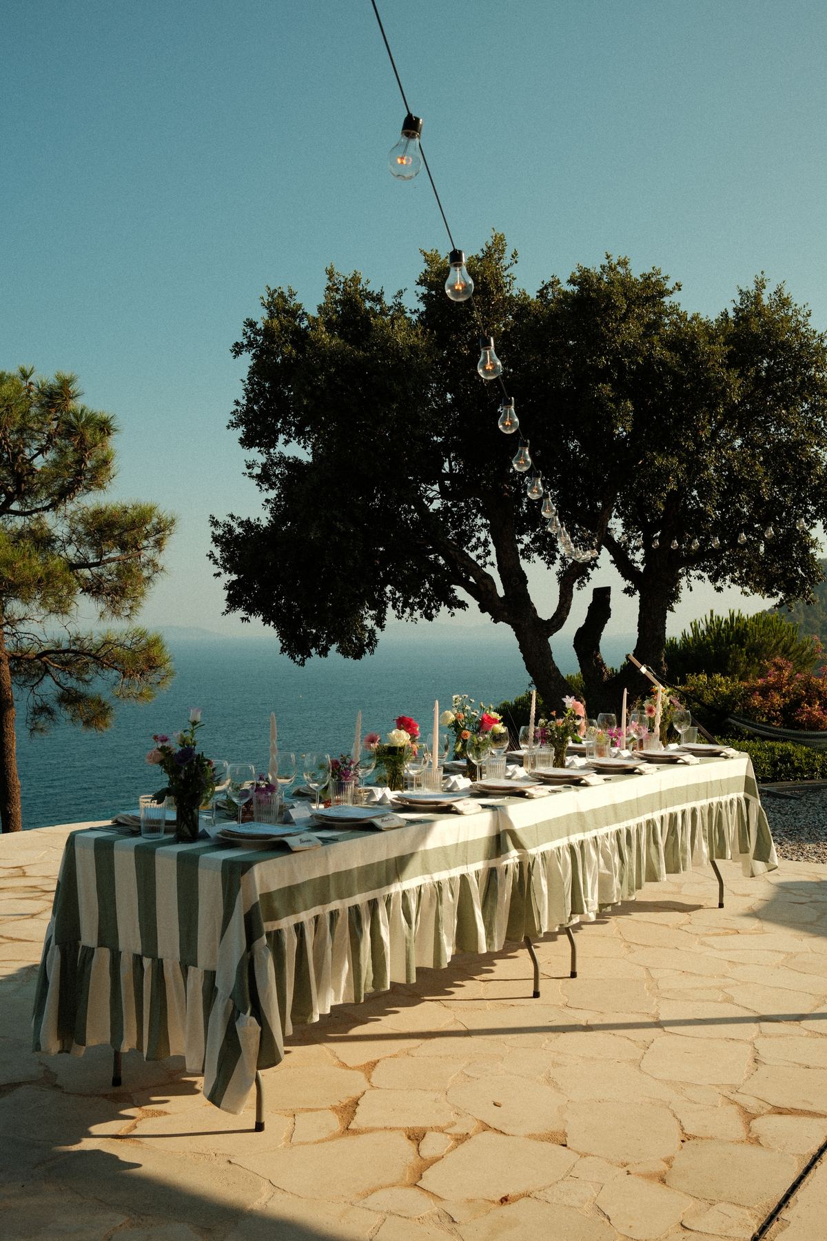 Long dinner table dressed with green-and-white striped linens and wildflowers, set beneath string lights in an olive tree overlooking the Mediterranean Sea at golden hour — from an 18th-birthday celebration